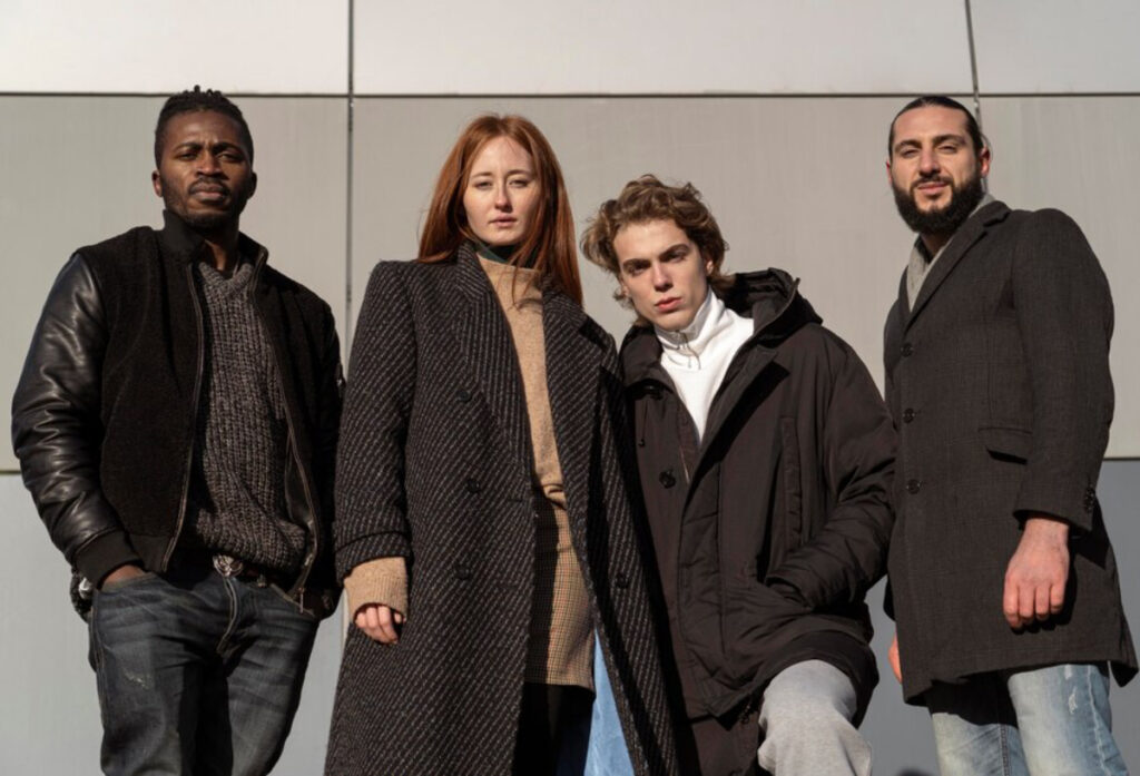 Four individuals stand together outdoors, dressed in stylish winter attire, against a modern architectural backdrop.