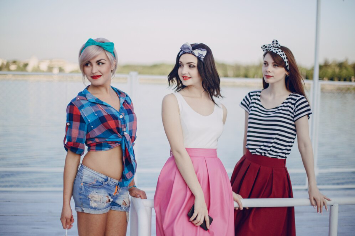 Three young women stand on a pier by a body of water, dressed in colorful outfits with hair accessories, enjoying a sunny day.