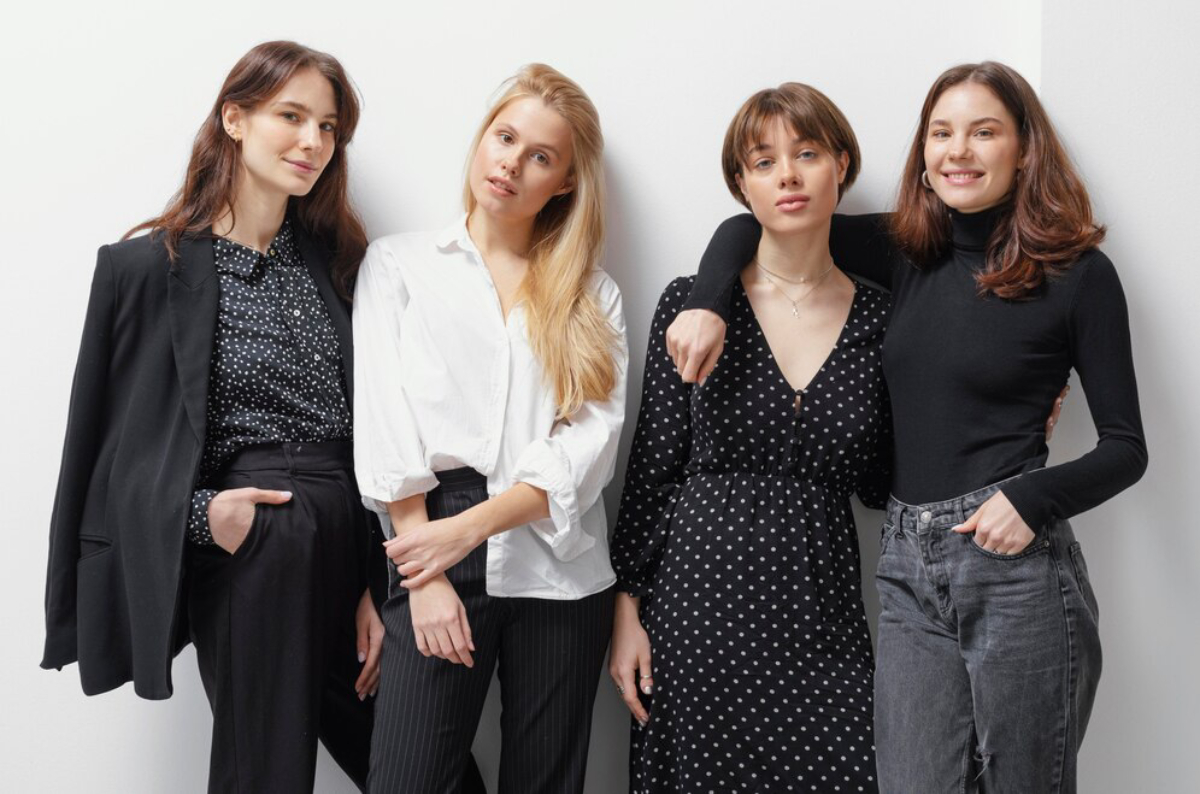 Four women stand together, dressed in stylish outfits featuring polka dots and bold colors, against a simple white background.