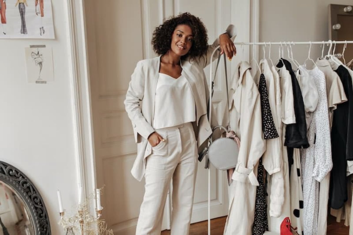 A stylish individual in a light suit and top stands next to a rack filled with an array of fashionable clothing in a chic room.