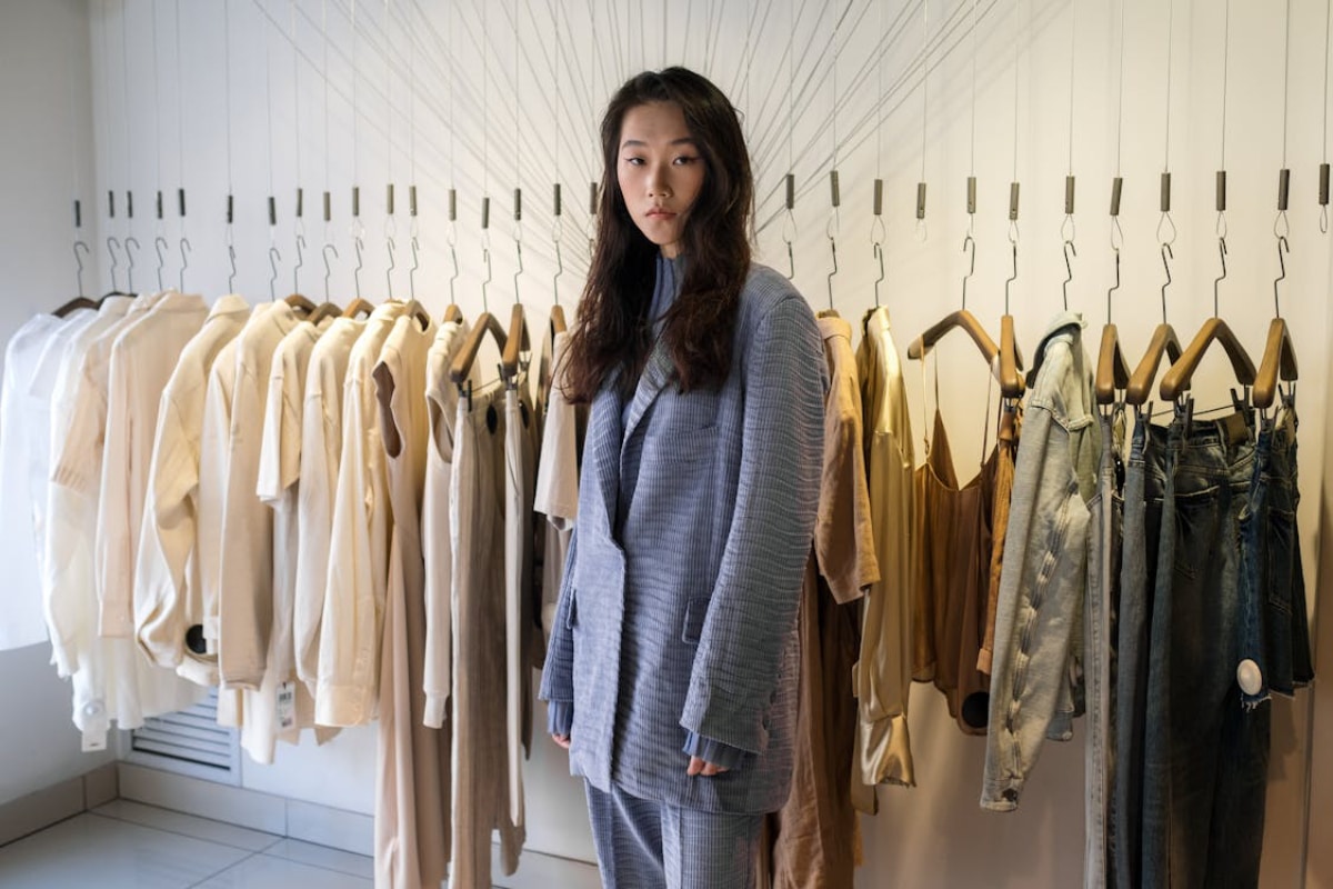 A person stands in a stylish shop surrounded by various clothing items on hangers, featuring neutral tones and contemporary designs.