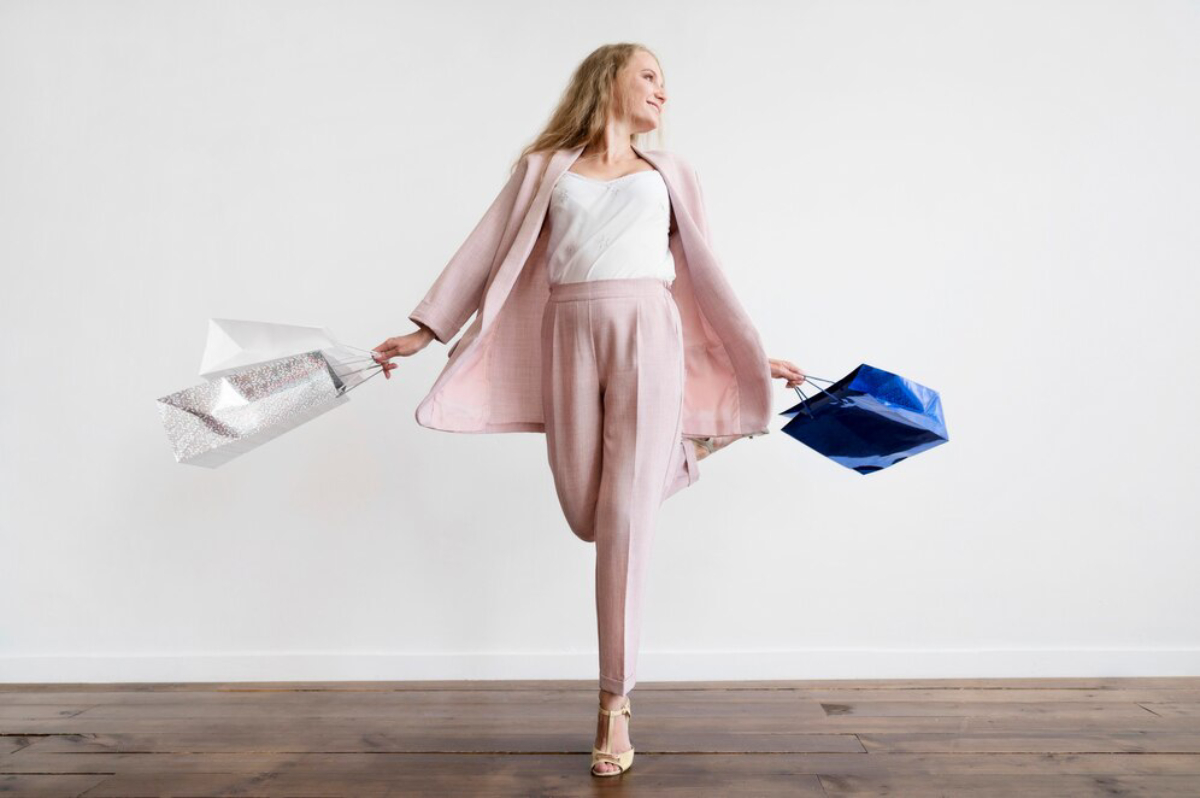 A woman in a chic pink suit holds silver and blue shopping bags, standing confidently against a white backdrop.