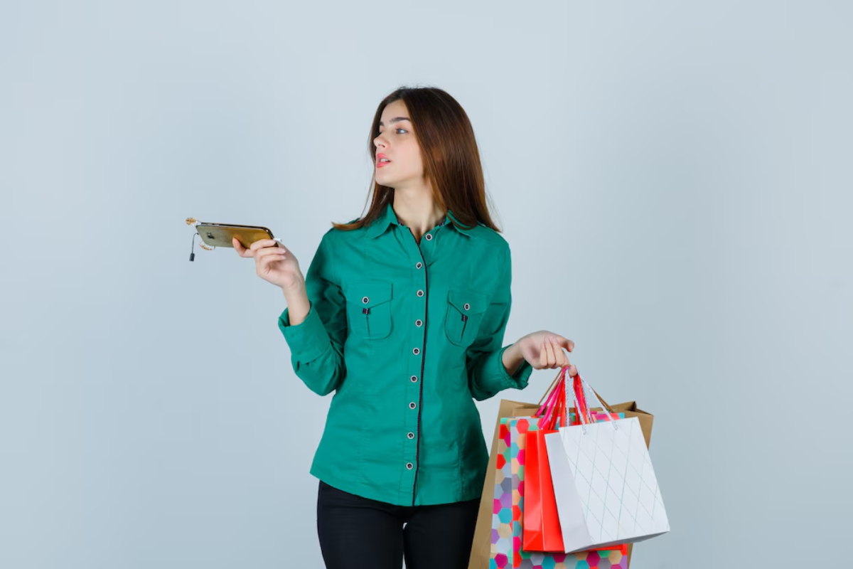 A woman in a green shirt holds a phone in one hand and several shopping bags in the other, standing against a light backdrop.