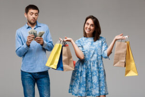 A man holds cash while a woman in a blue floral dress showcases colorful shopping bags, against a neutral gray background.