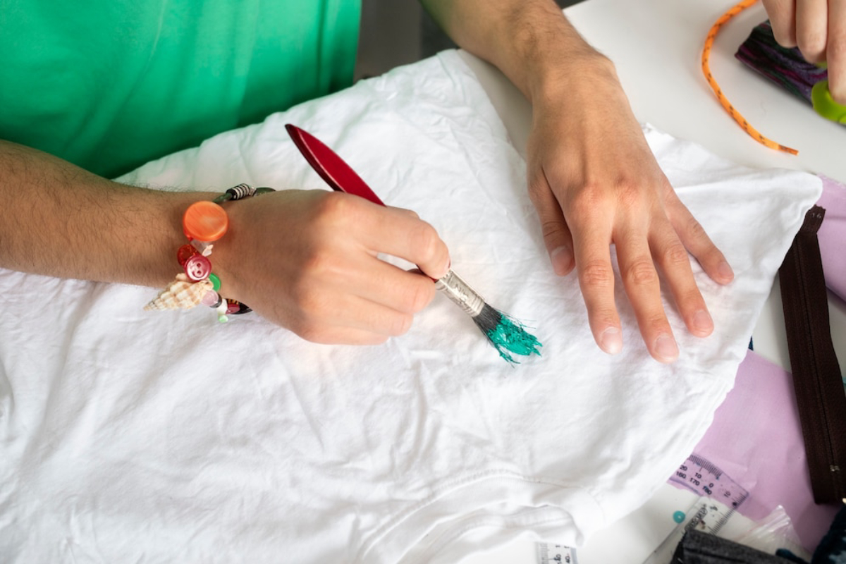 Person painting a white T-shirt with turquoise paint using a brush, surrounded by crafting materials.