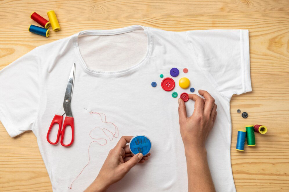 Hands arranging colourful buttons on a white T-shirt with sewing supplies on a wooden table.