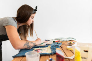 An artist paints on denim jeans at a wooden table, surrounded by various art supplies and tools. Creativity in action!