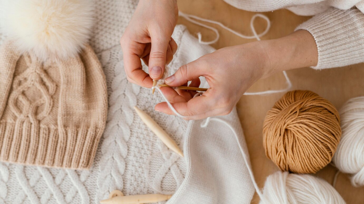 A pair of hands skillfully knitting with bamboo needles, surrounded by yarn, a cozy hat, and a textured sweater on a wooden table.