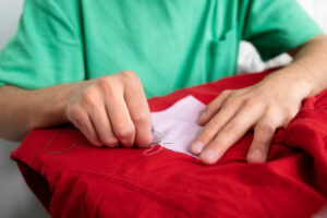 A person in a green shirt is sewing a white patch onto a red garment, focused on the task with thread in hand.