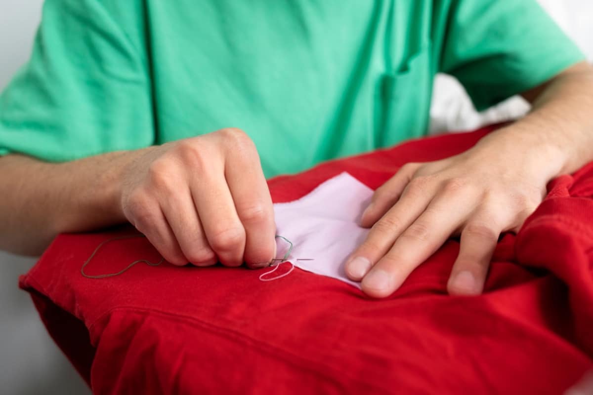 A person in a green shirt is sewing a white patch onto a red garment, focused on the task with thread in hand.