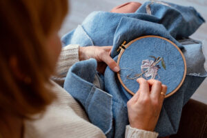 A woman hand-embroiders a floral design onto denim fabric using a wooden hoop, showcasing her craftsmanship and creativity.