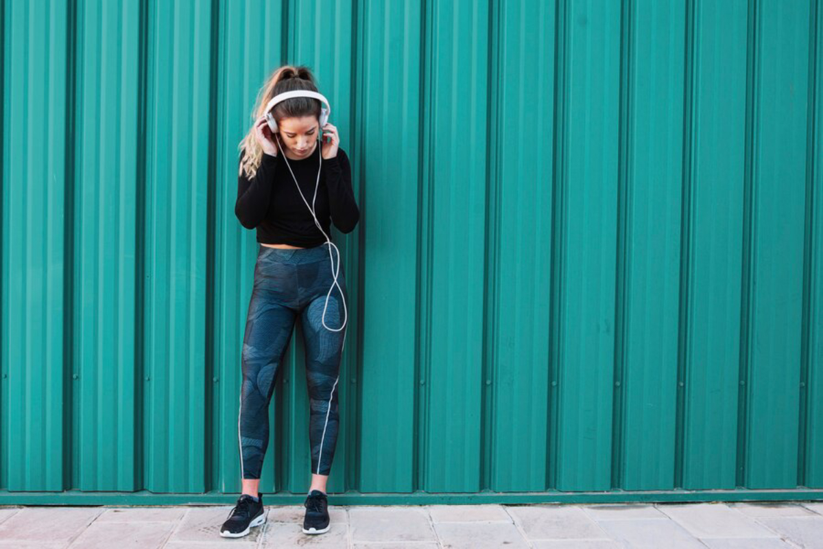 A person stands against a teal metal wall, wearing a black long-sleeve top and stylish patterned leggings, listening to headphones.