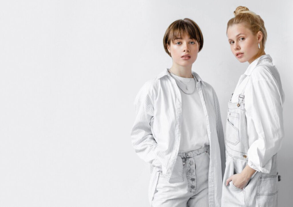 Two stylish individuals stand side by side, wearing coordinated outfits in shades of white and light gray against a minimalist background.