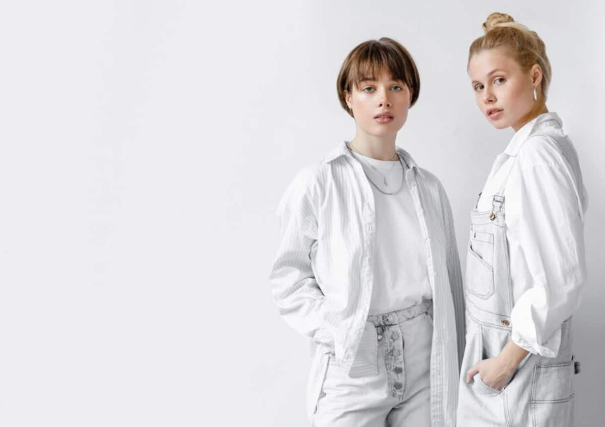 Two stylish individuals stand side by side, wearing coordinated outfits in shades of white and light gray against a minimalist background.