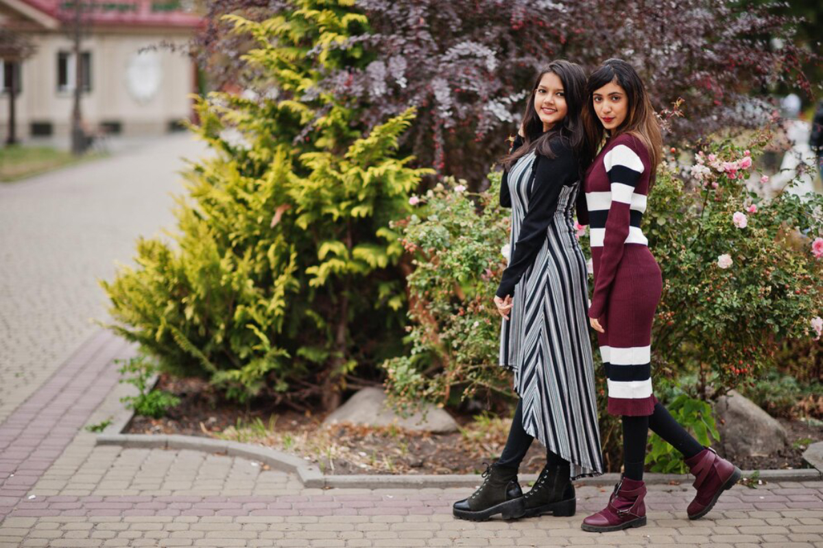 Two women stand side by side in a garden, one in a striped dress and the other in a maroon striped sweater, surrounded by greenery.
