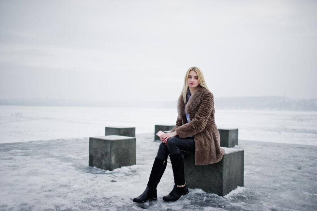A woman sits on a concrete block by a frozen lake, dressed in a brown fur coat, black pants, and tall black boots under a cloudy sky.