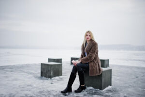 A woman sits on a concrete block by a frozen lake, dressed in a brown fur coat, black pants, and tall black boots under a cloudy sky.