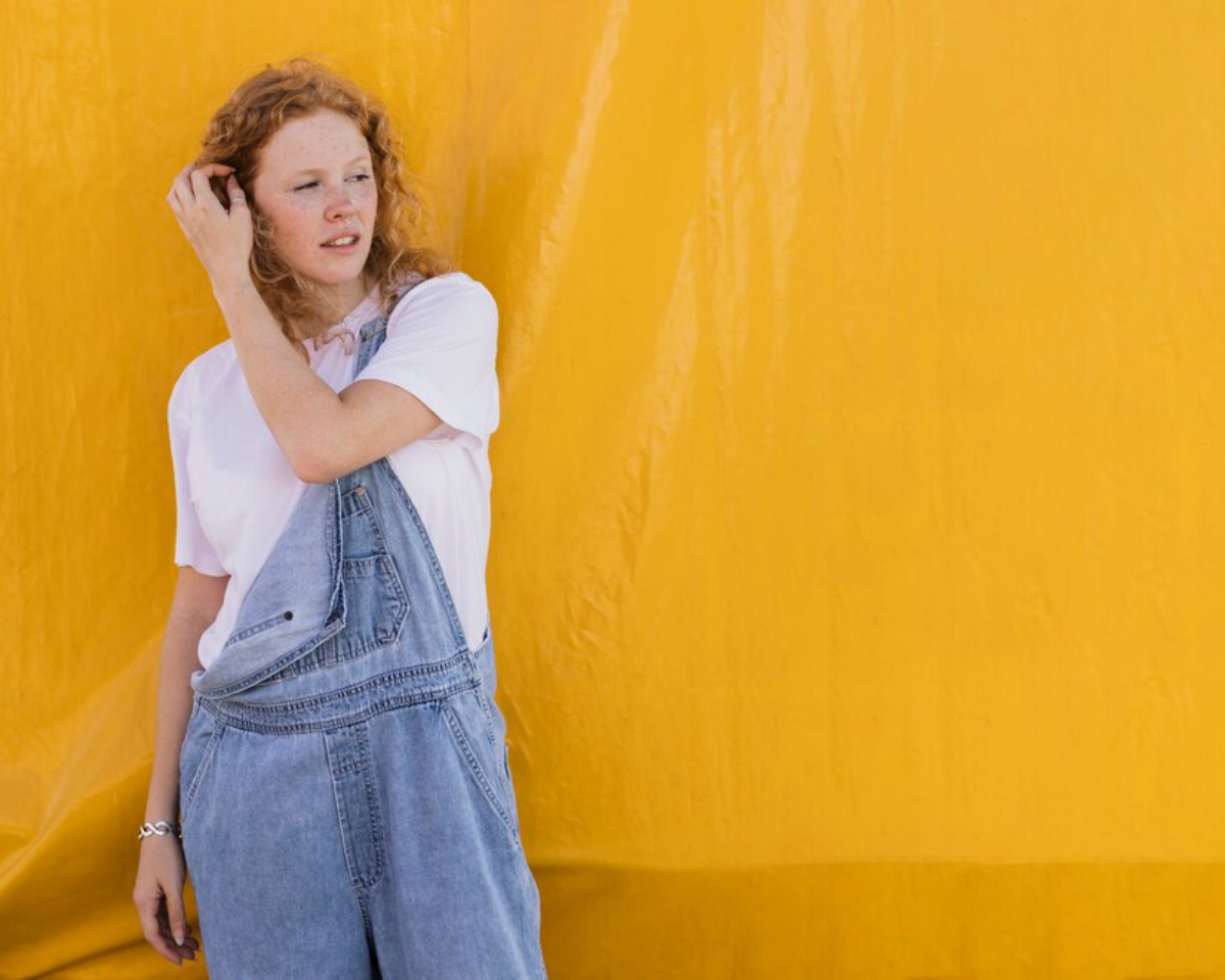 A person stands against a vibrant yellow backdrop, wearing a white t-shirt and denim overalls, casually touching their hair.
