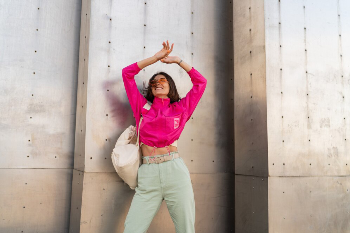 A person poses confidently against a metallic wall, wearing a vibrant pink cropped shirt and light green pants, with a beige bag.