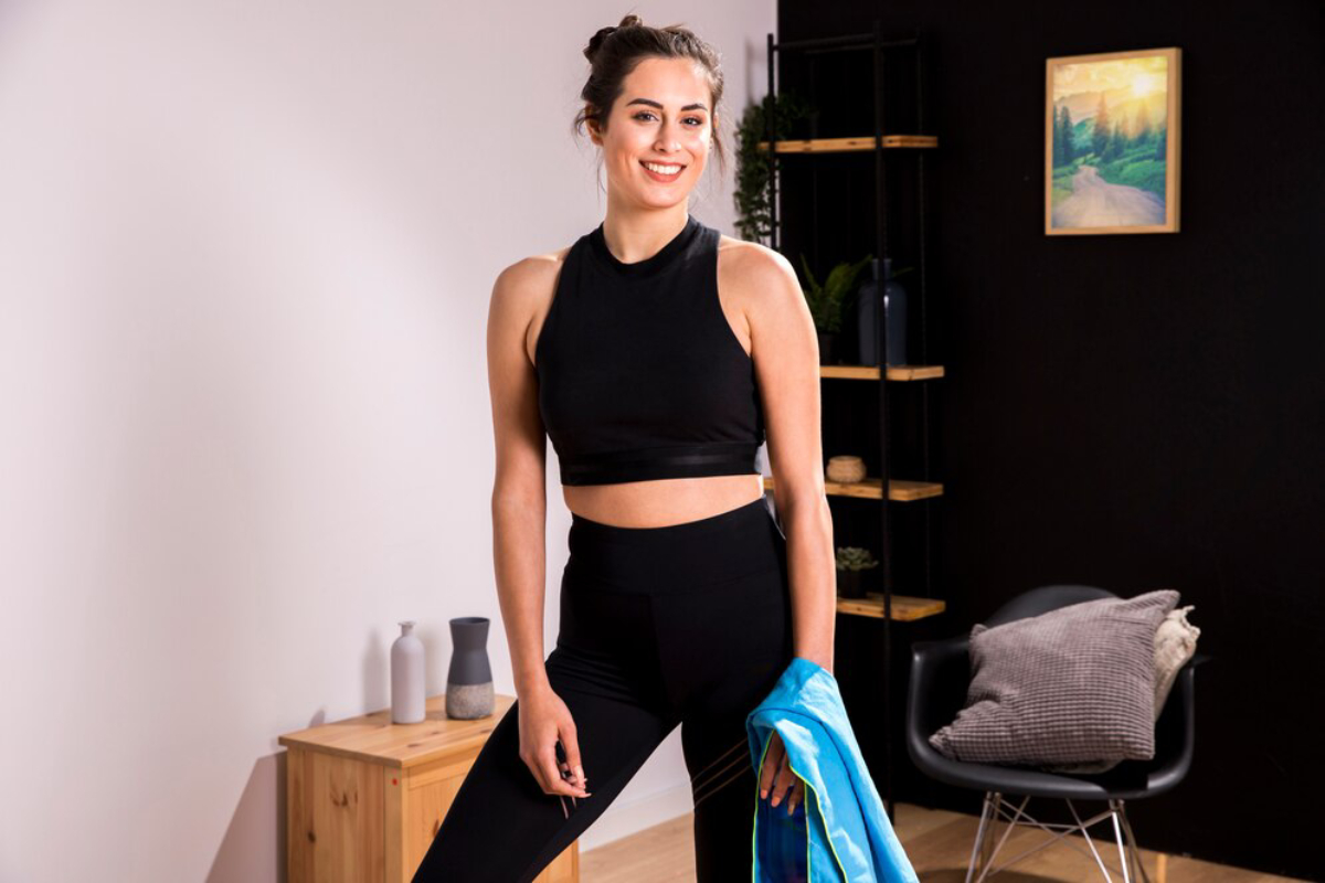 A person in athletic wear stands confidently indoors, holding a blue jacket, with modern decor and plants in the background.