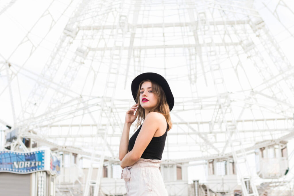 A woman in a black hat poses confidently in front of an intricate white structure, embodying a stylish urban vibe.