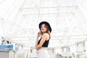 A woman in a black hat poses confidently in front of an intricate white structure, embodying a stylish urban vibe.