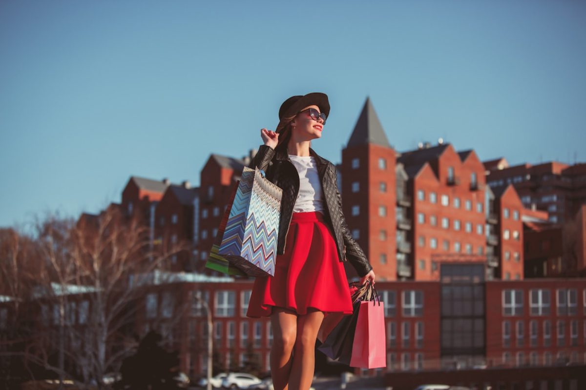 A woman in a red skirt and black jacket smiles while holding colorful shopping bags against a backdrop of modern buildings.