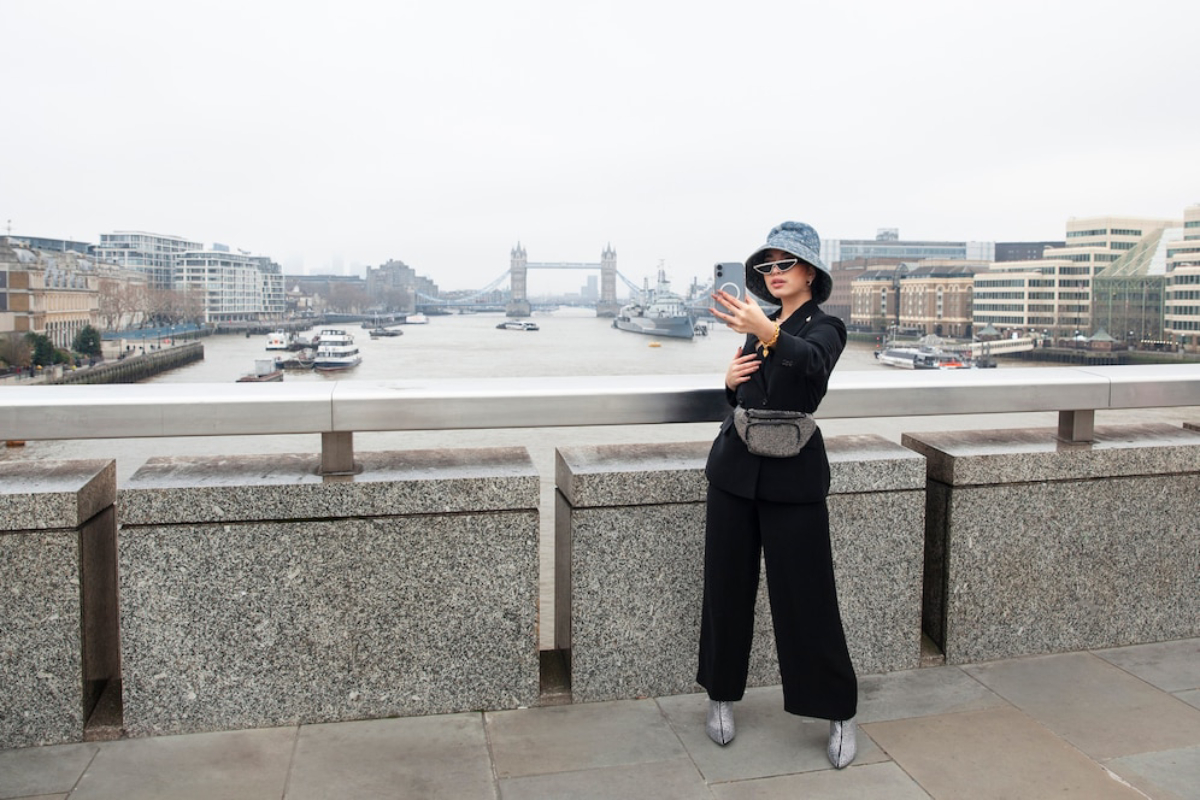 A person in stylish black attire poses for a selfie in front of the River Thames, with Tower Bridge in the background on a cloudy day.