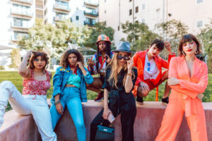 A group of six fashionable friends poses playfully outdoors, dressed in vibrant styles, with modern apartment buildings in the background.