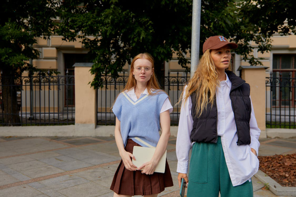 Two young women stand outdoors, one wearing a blue vest over a white shirt and the other in a black puffer vest and green pants.
