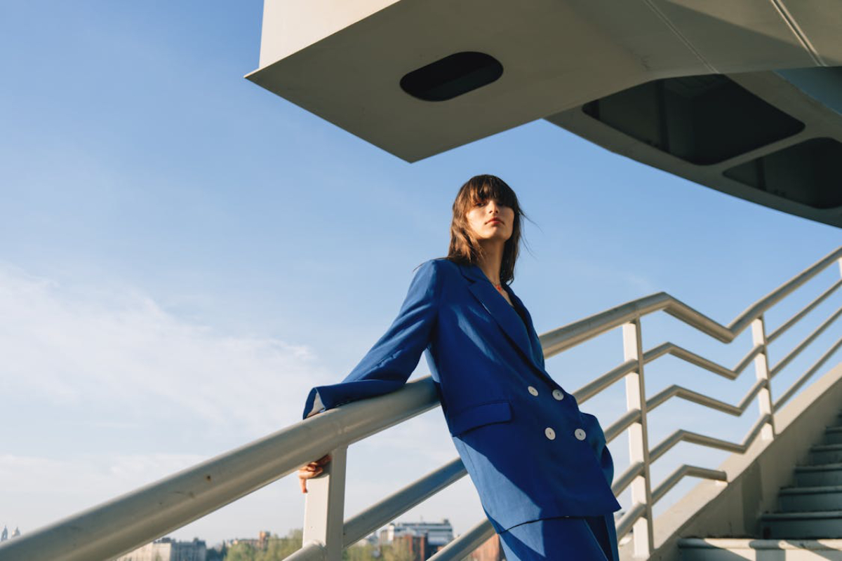 A person in a blue suit leans casually against a railing, with a modern architectural structure and clear blue sky in the background.
