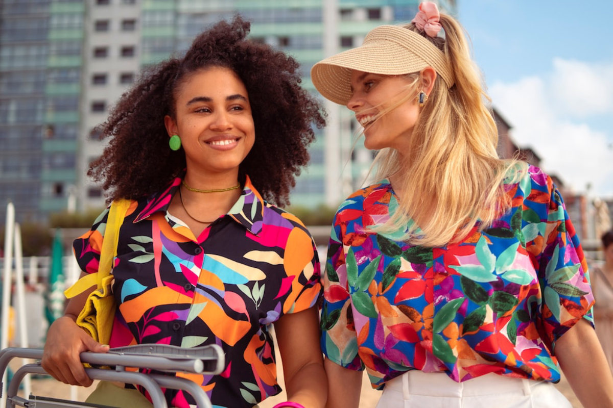 Two women in vibrant, patterned shirts stroll along a sandy beach, with a modern cityscape in the background.