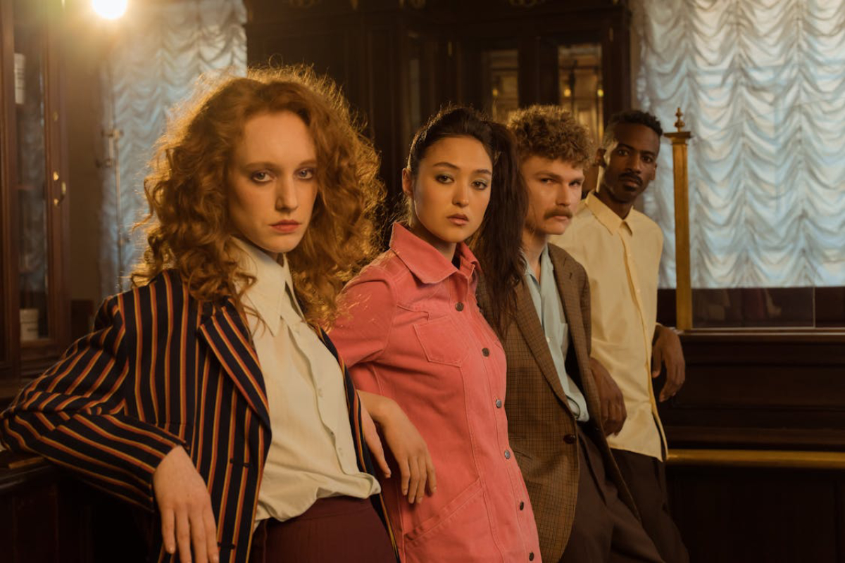 Four stylish individuals pose together, showcasing diverse fashion choices and hair styles in a warm, vintage interior setting.