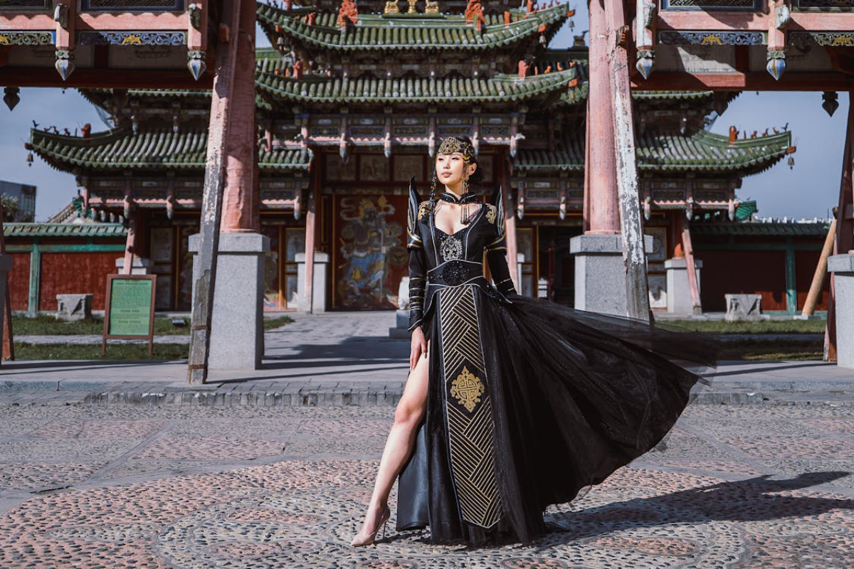 A person in a stunning black gown with intricate gold detailing poses gracefully in front of ornate, traditional architecture.