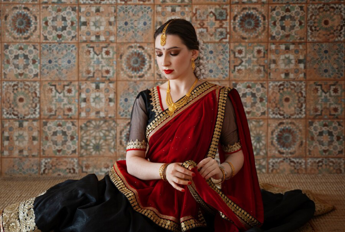 A person dressed in a red and black traditional outfit, adorned with gold embellishments, poses against a colorful tiled background.