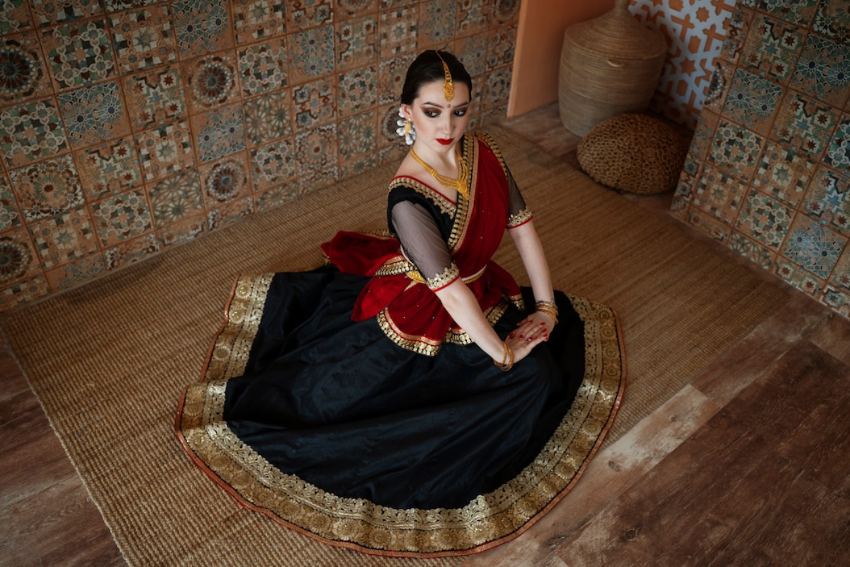A person dressed in a black and red traditional outfit poses gracefully on a woven mat, with patterned walls in the background.