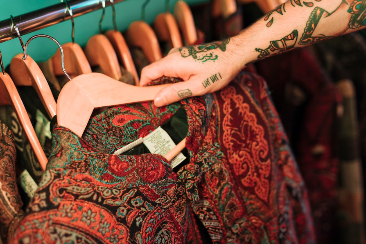 A tattooed hand selects a richly patterned garment from a rack of vintage clothing hangers. Vibrant colors and intricate designs are visible.