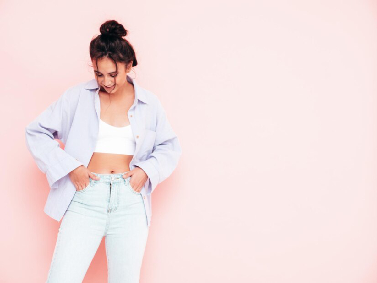A person in a light purple oversized shirt with a white crop top and high-waisted jeans poses against a soft pink background.