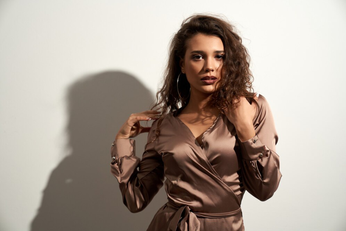 A woman poses confidently in a silky brown wrap dress, showcasing long sleeves and a stylish silhouette against a simple white background.