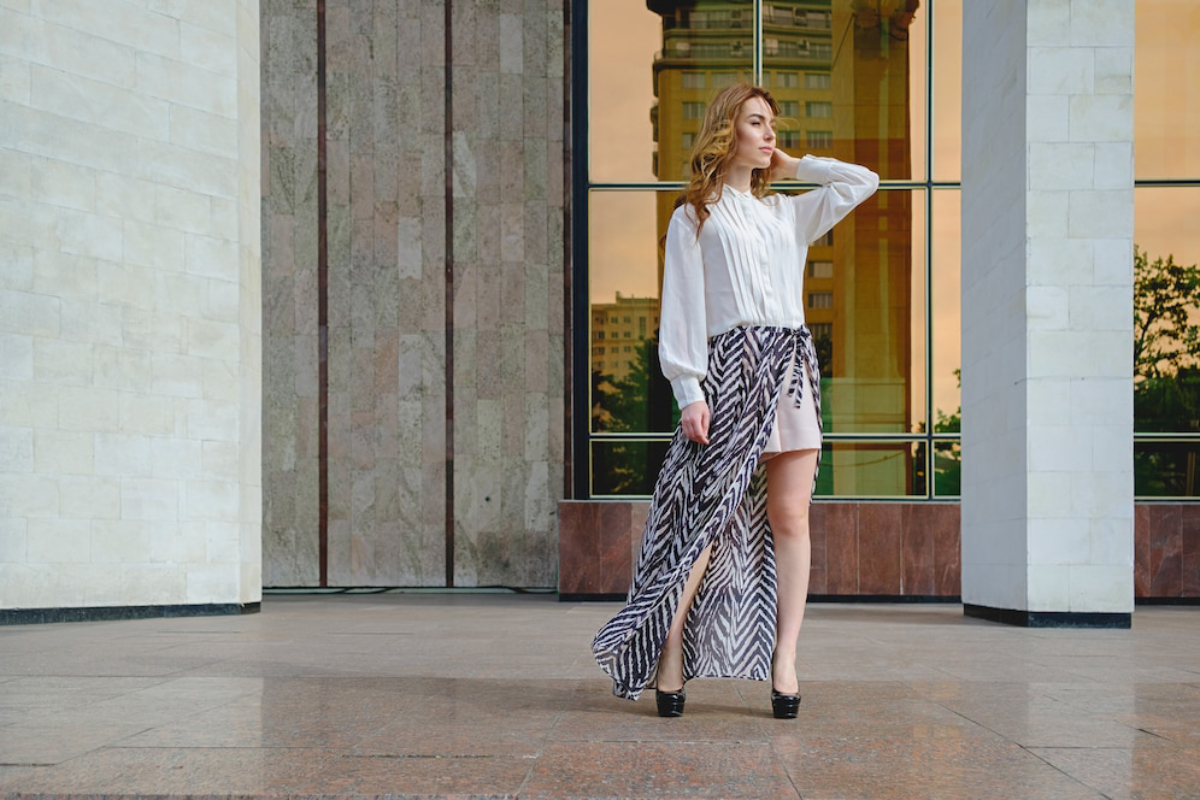 A woman in a light blouse and a flowing, patterned skirt stands confidently on a polished stone surface against a modern building backdrop.