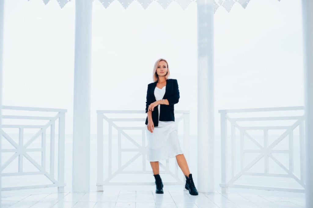 A stylish individual poses confidently in a white dress and black jacket, set against a bright, airy backdrop with white railings.