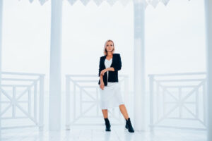 A stylish individual poses confidently in a white dress and black jacket, set against a bright, airy backdrop with white railings.