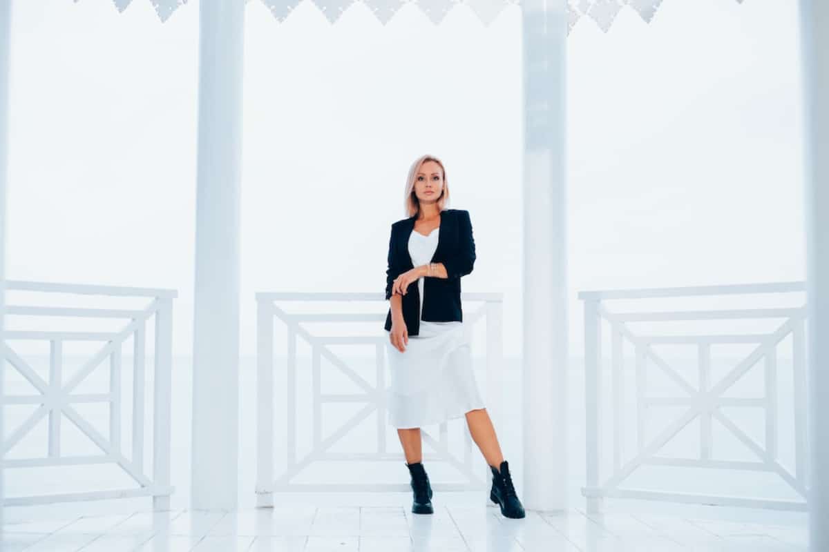 A stylish individual poses confidently in a white dress and black jacket, set against a bright, airy backdrop with white railings.