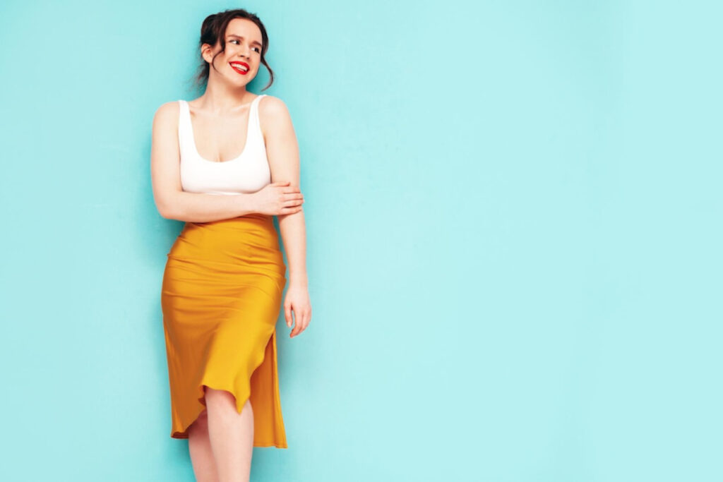 A woman stands against a bright turquoise wall, wearing a fitted white tank top and a flowing mustard yellow skirt.