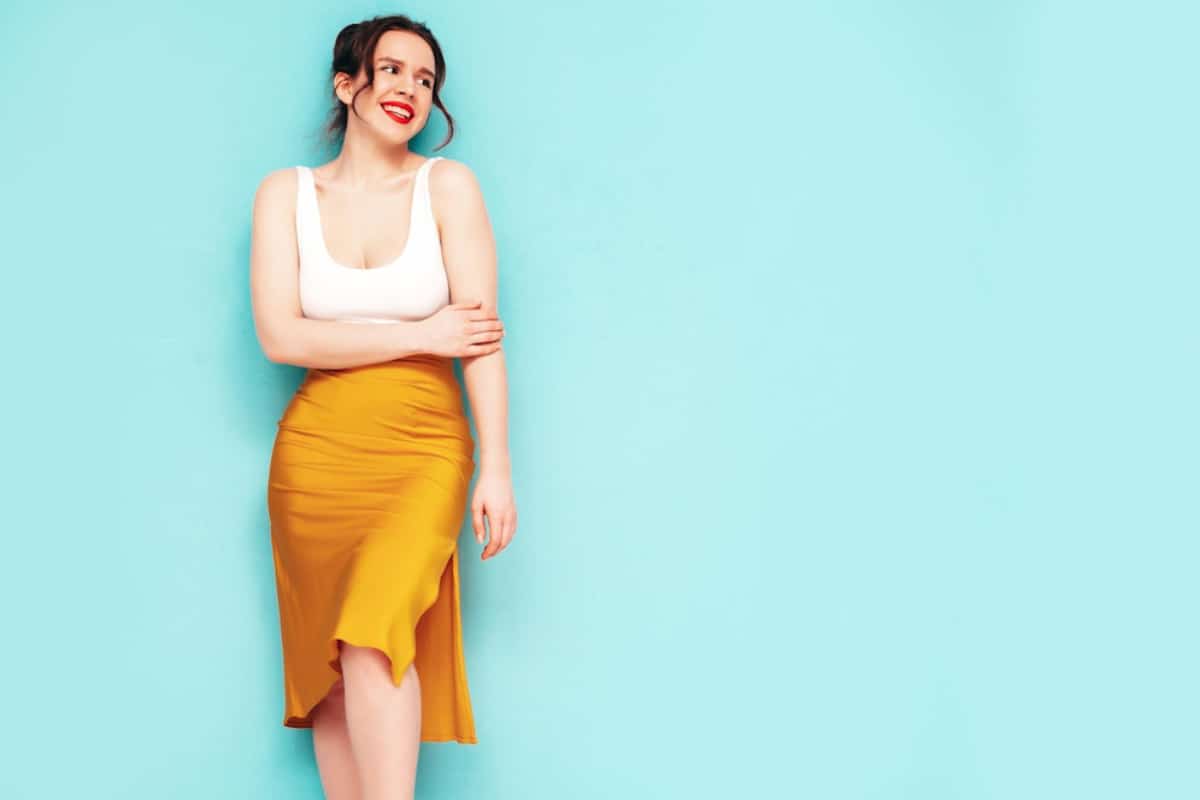 A woman stands against a bright turquoise wall, wearing a fitted white tank top and a flowing mustard yellow skirt.
