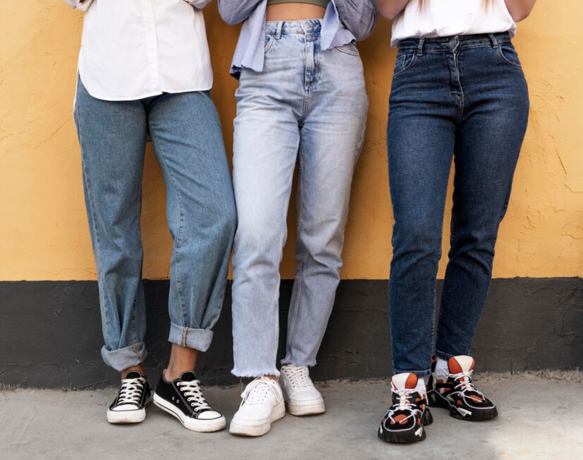 Three women stand side by side against a yellow wall, showcasing a variety of denim styles and trendy sneakers.