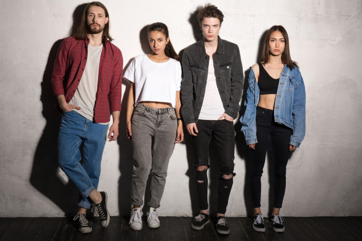 Four stylish young adults stand against a textured wall, showcasing trendy outfits featuring denim, cropped tops, and casual sneakers.