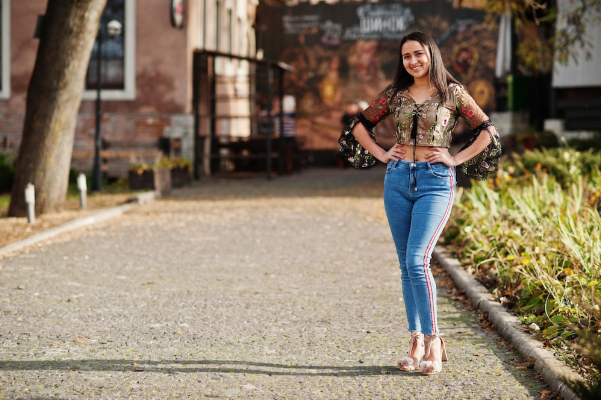A stylish person poses on a scenic cobblestone path, wearing a floral top and skinny jeans, with greenery and rustic buildings in the background.