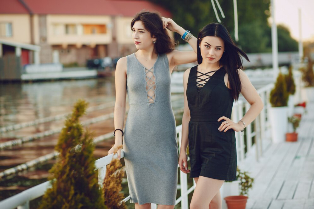 Two women pose together by a waterfront, wearing stylish outfits: one in a gray dress and the other in a black romper.