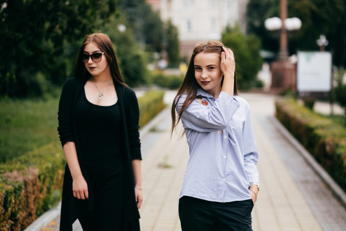 Two women stroll down a leafy pathway, one adjusting her hair and the other gazing ahead, surrounded by greenery and city buildings.